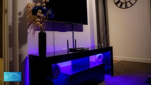 A modern living room with a flat-screen TV mounted on a black entertainment unit, a decorative vase with dried flowers, and a wall clock.