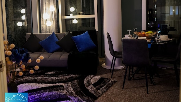 A modern living room with a sofa, dining table, and chairs. There are large windows with curtains and a view of the city.