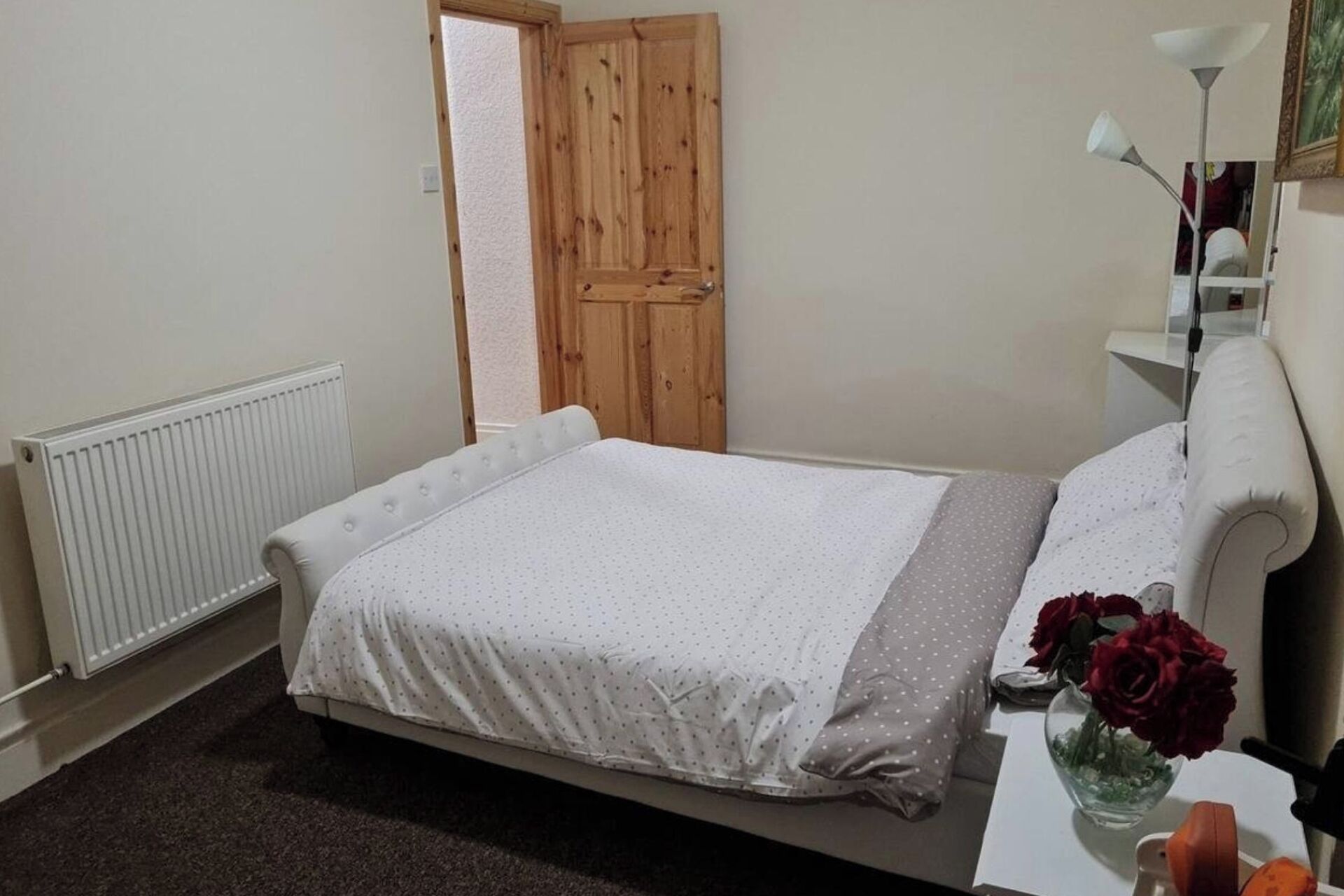 A bedroom with a bed, a radiator, a wooden door, a white bedside table, and a vase with flowers.