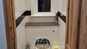 A bathroom with white tiles, a window, a toilet with a decorative cover, and a small shelf with toiletries.