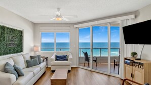 A living room with a sofa, coffee table, and a television. There is a balcony with a view of the ocean. - Cozy Pet Friendly Beachfront Retreat On Top Floor (Orange Beach)