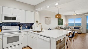 A modern kitchen with white cabinetry, a built-in microwave, and a dishwasher. There is a large island with a sink and a dining area with a view of the ocean. - Cozy Pet Friendly Beachfront Retreat On Top Floor (Orange Beach)