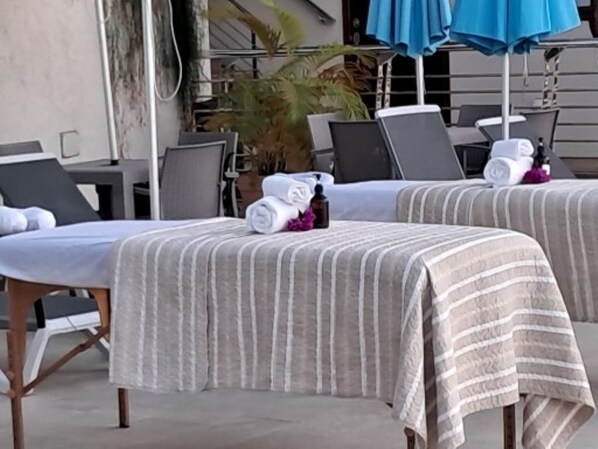 Dos camillas de spa con toallas a rayas, botellas, una planta en maceta y sombrillas exteriores.