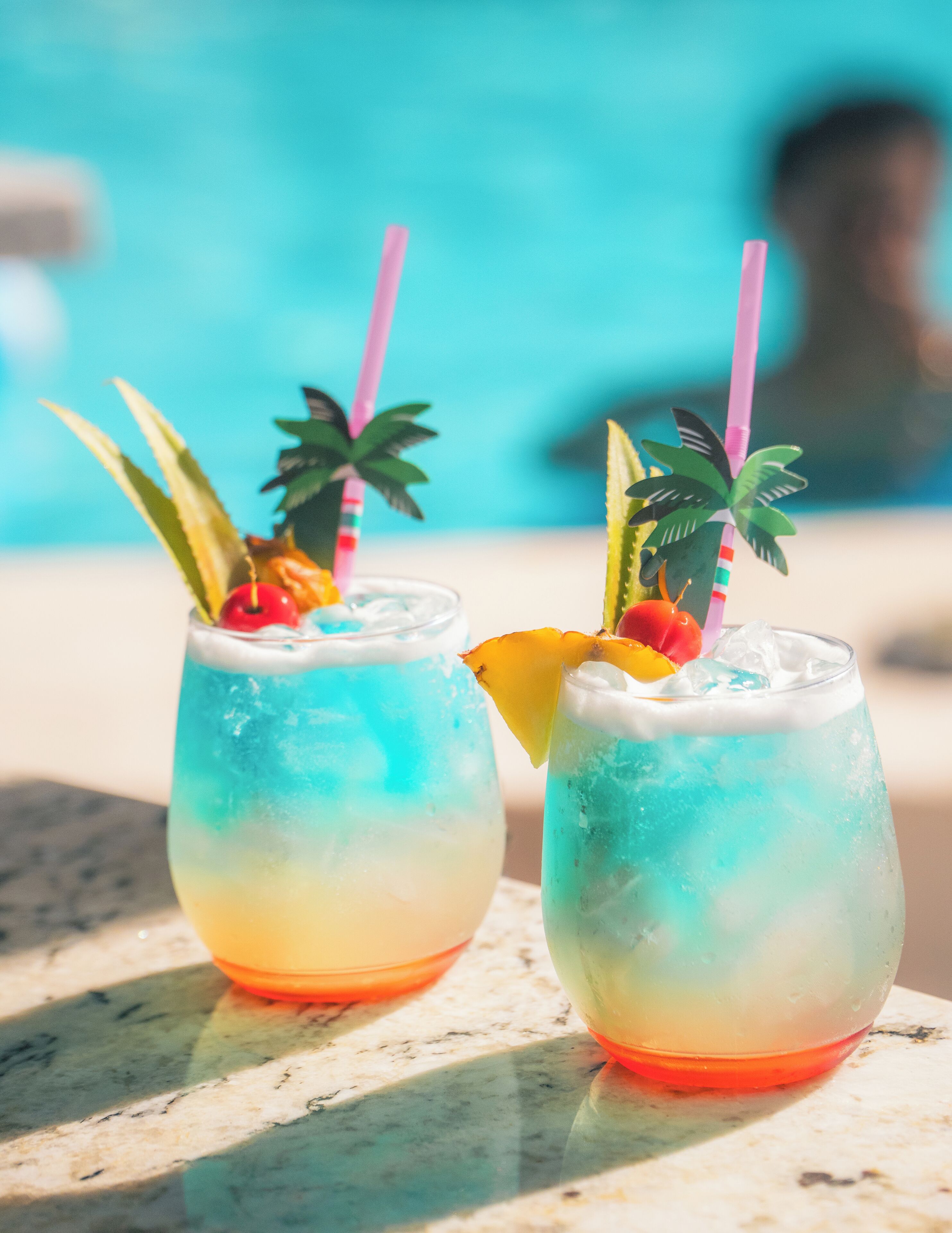 Two cocktails with tropical garnishes and straws, placed on a table near a pool.