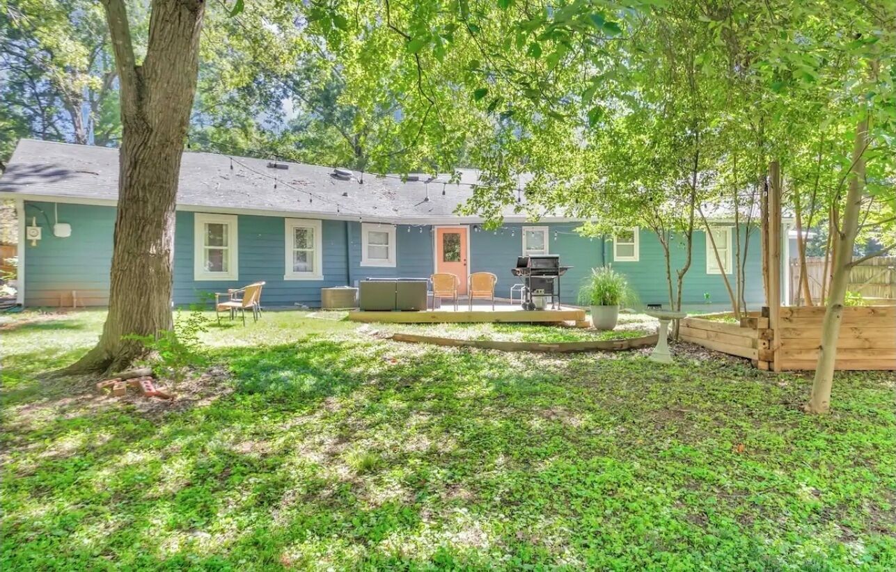 A house with a backyard featuring a barbecue grill, outdoor seating, and a large tree.
