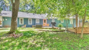 A house with a backyard featuring a barbecue grill, outdoor seating, and a large tree.