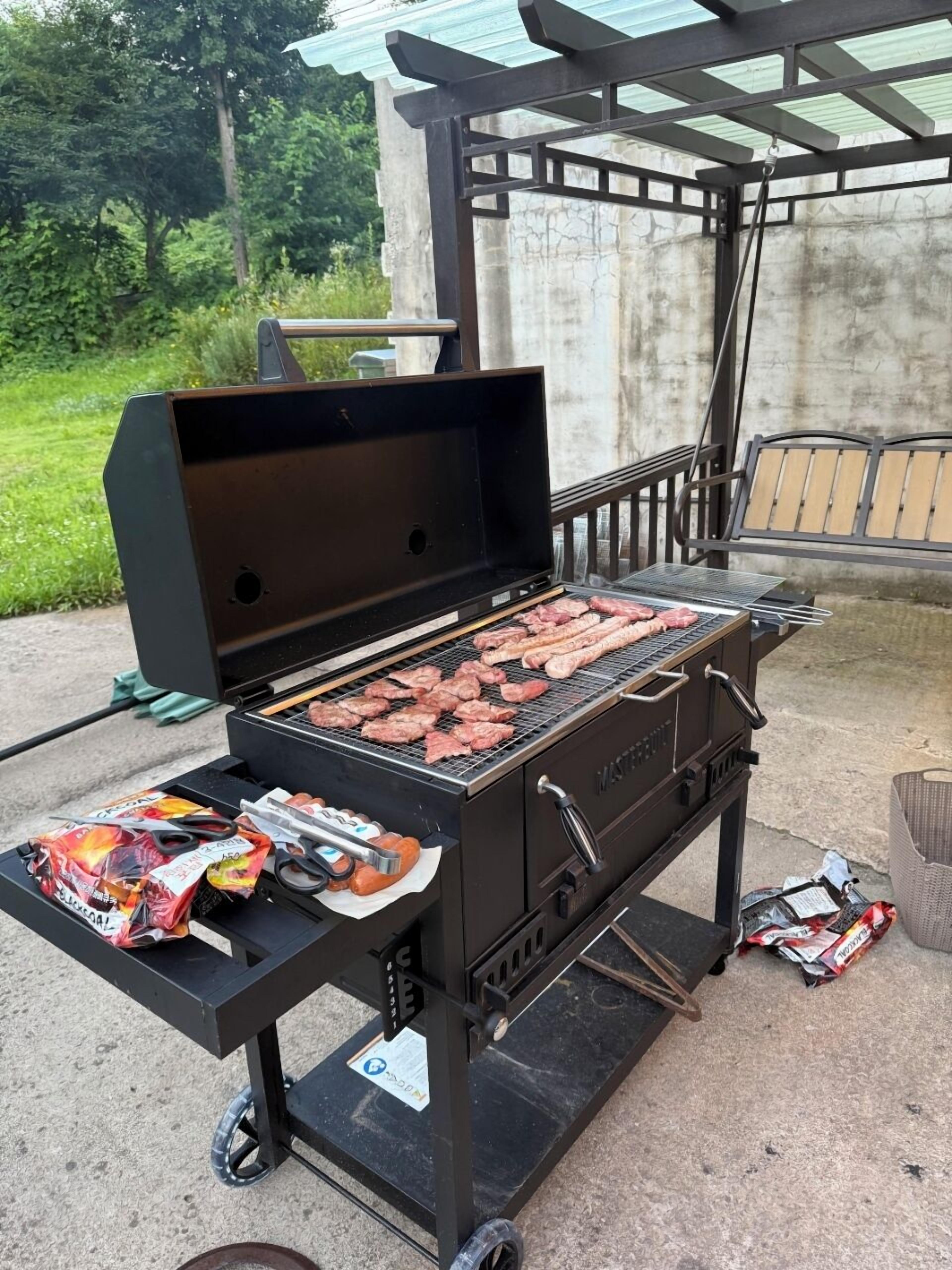 Una parrilla portátil con varios tipos de carne asándose, un saco de carbón vegetal y un par de tijeras sobre una mesa.