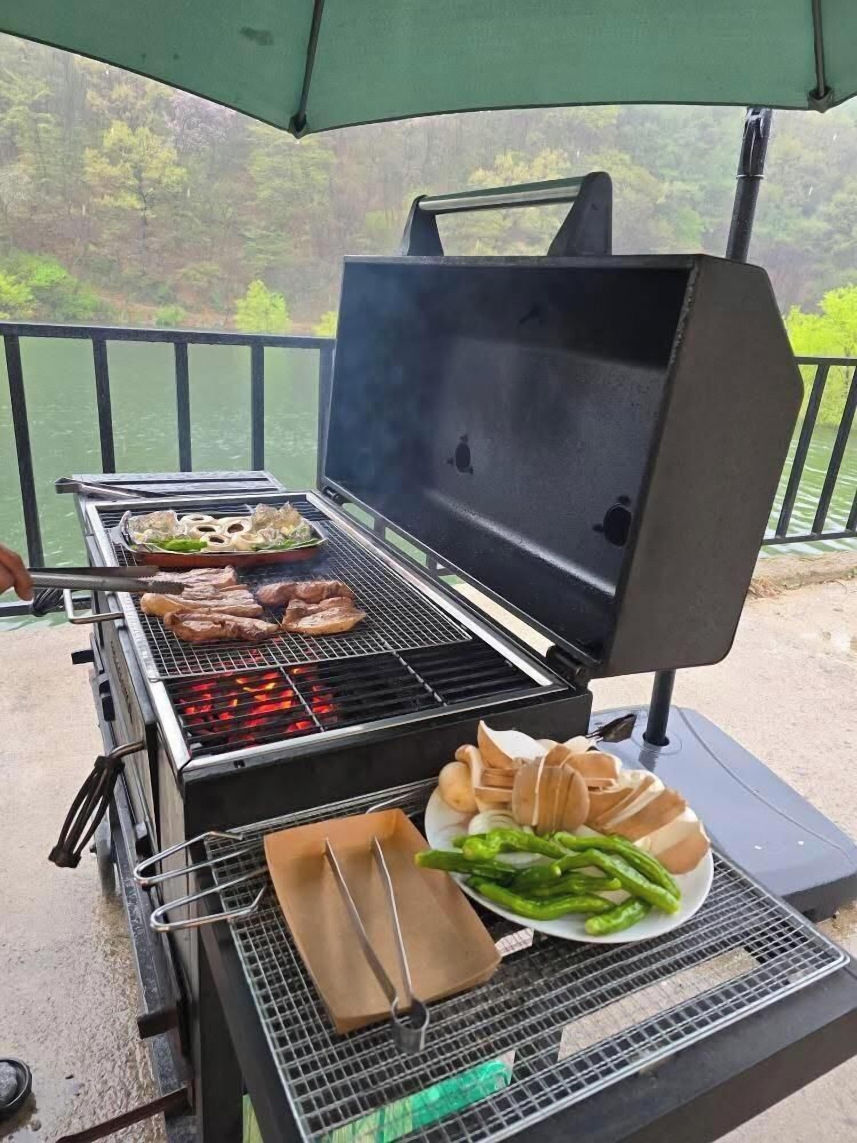 A portable grill with food being cooked, a plate with green vegetables, and a person grilling meat.
