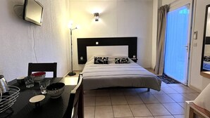 A hotel room with a bed, a desk with a chair, a small table with bowls, a lamp, and a TV mounted on the wall.