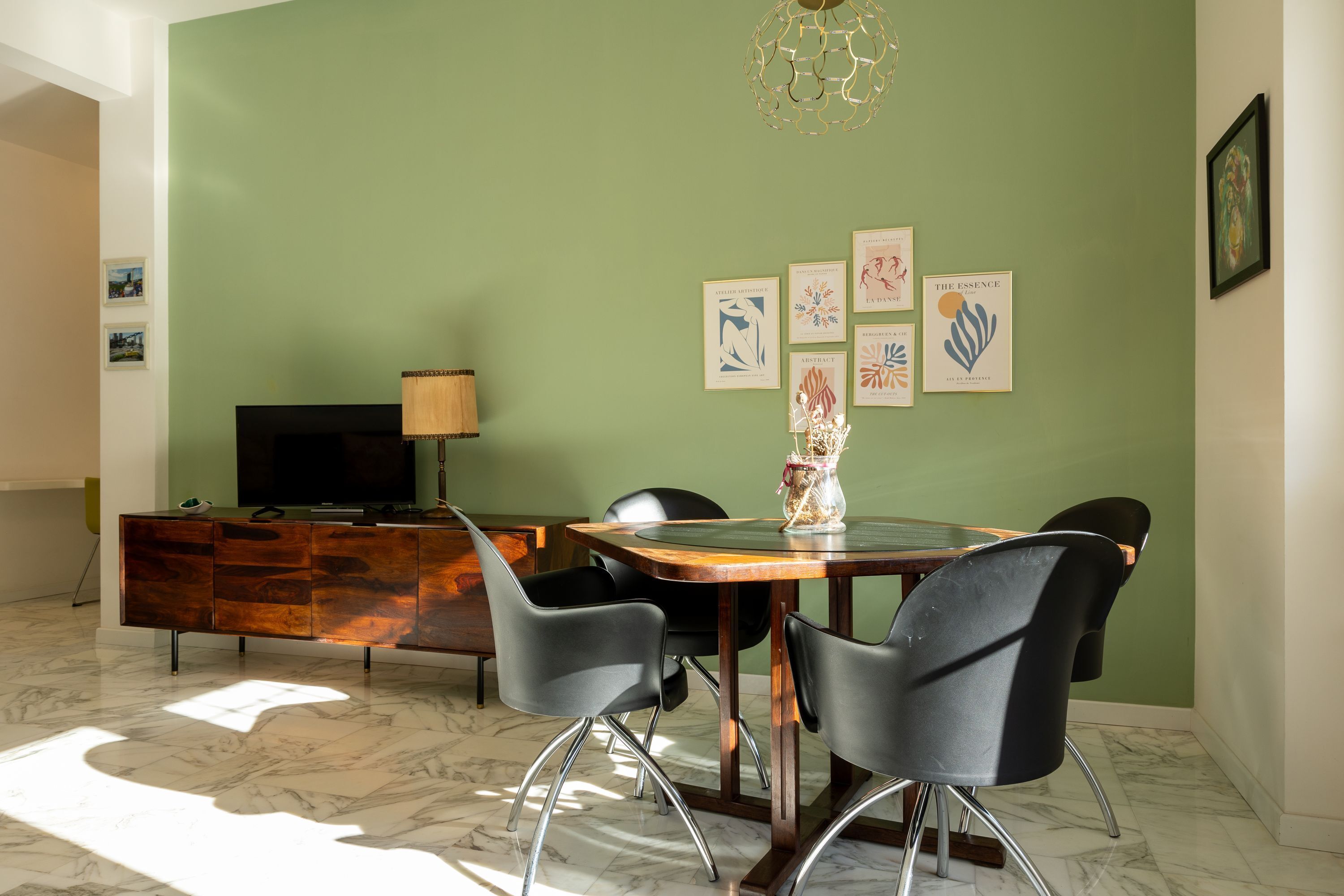Un coin repas avec un sol en marbre, une table en bois, des chaises noires, un téléviseur posé sur un meuble en bois et des œuvres d’art encadrées accrochées au mur.