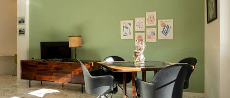 Un coin repas avec un sol en marbre, une table en bois, des chaises noires, un téléviseur posé sur un meuble en bois et des œuvres d’art encadrées accrochées au mur.