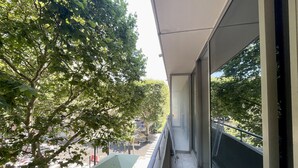 Un balcon donnant sur une rue bordée d’arbres, avec des sièges pour l’extérieur et une porte vitrée.