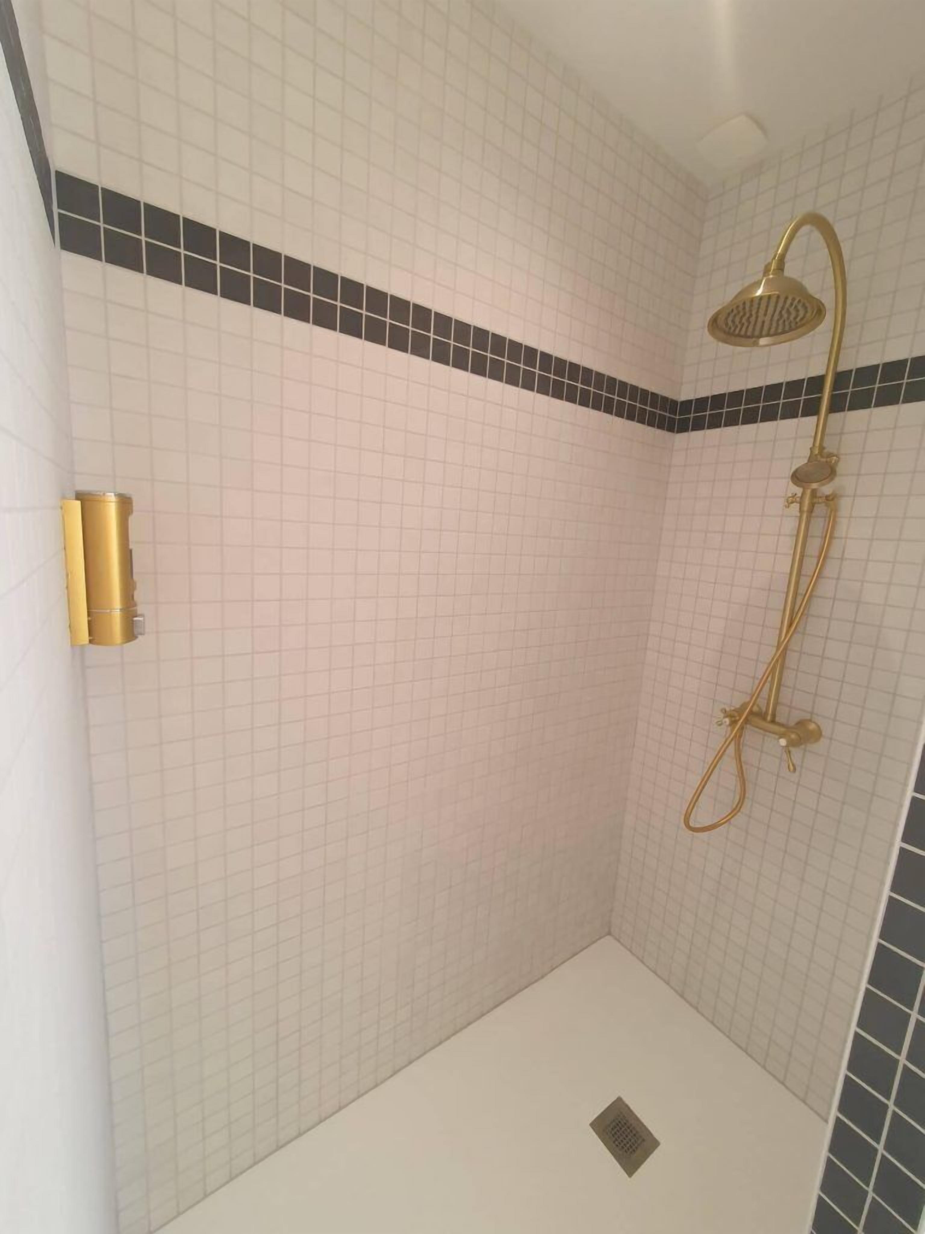 A small, tiled shower room with a gold showerhead and a wall-mounted light fixture.