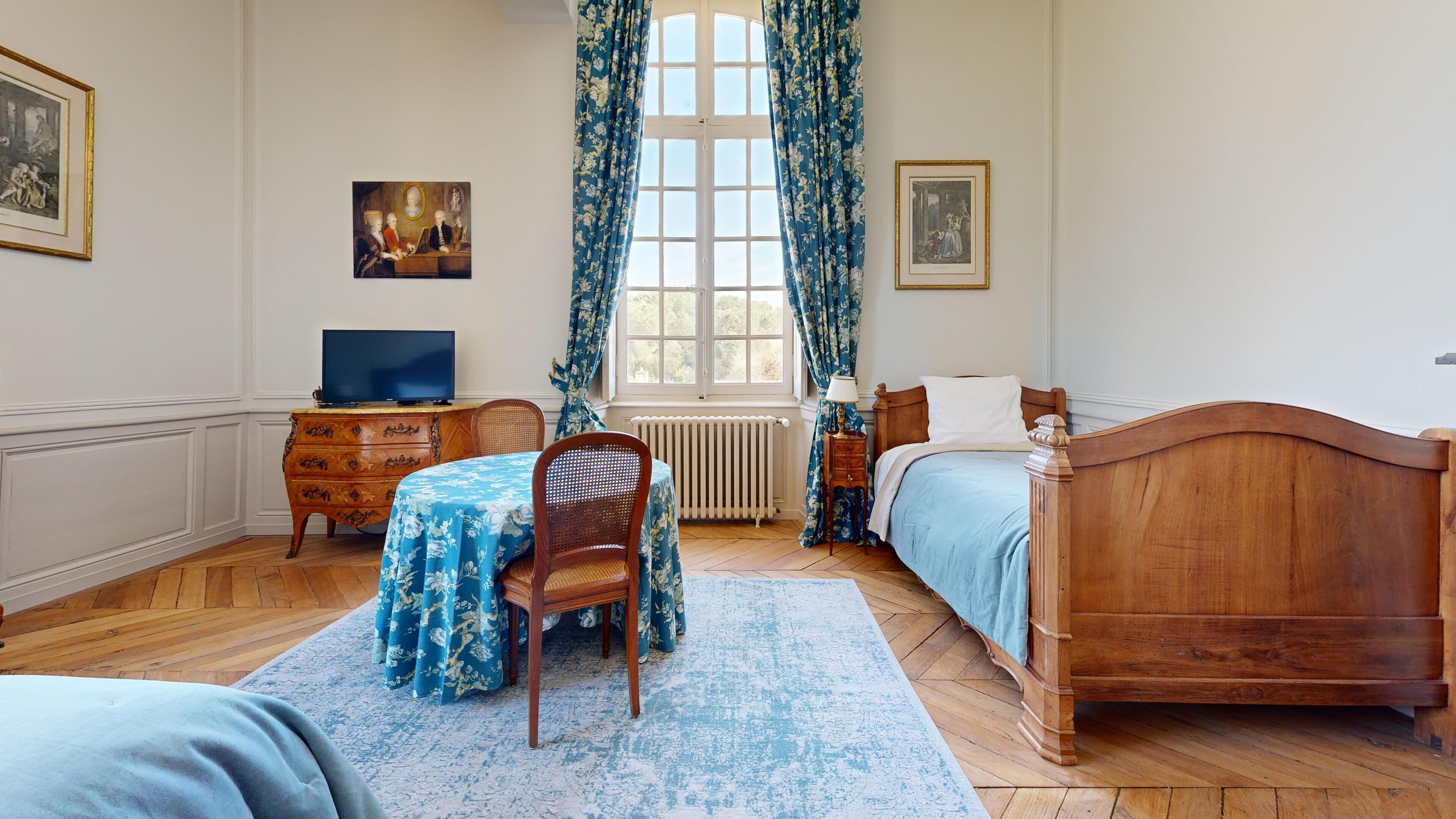 Un dormitorio con una cama de madera, un escritorio con televisor, una silla, una mesa con mantel floreado y dos cuadros en la pared.