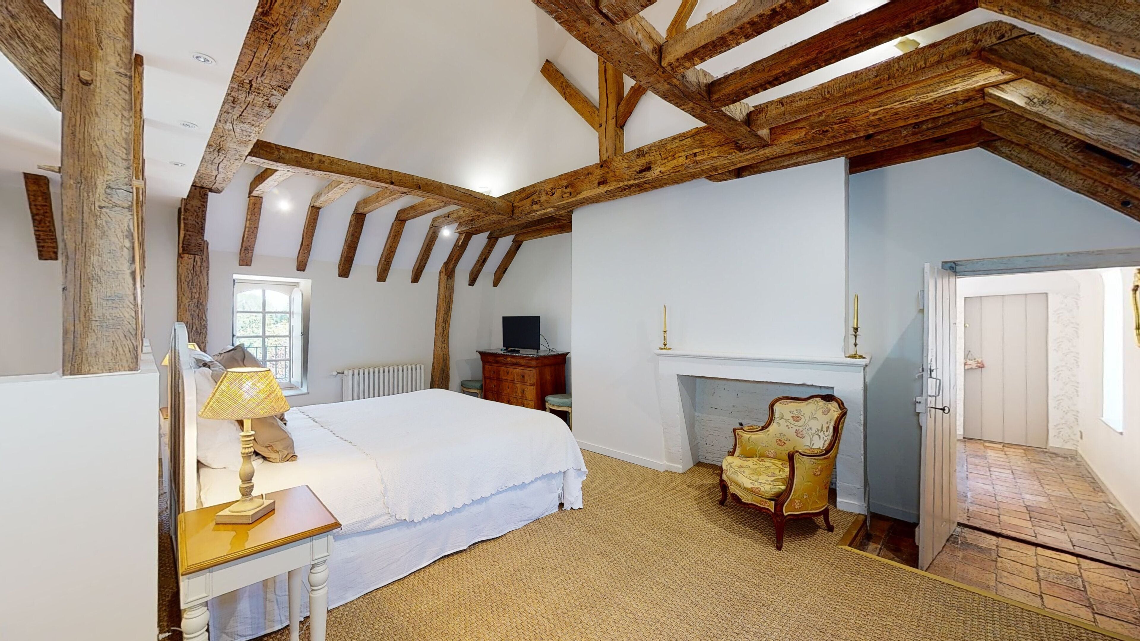 Un dormitorio con vigas de madera a la vista, una cama, un aparador, una silla, una chimenea y una puerta que da a otra habitación.