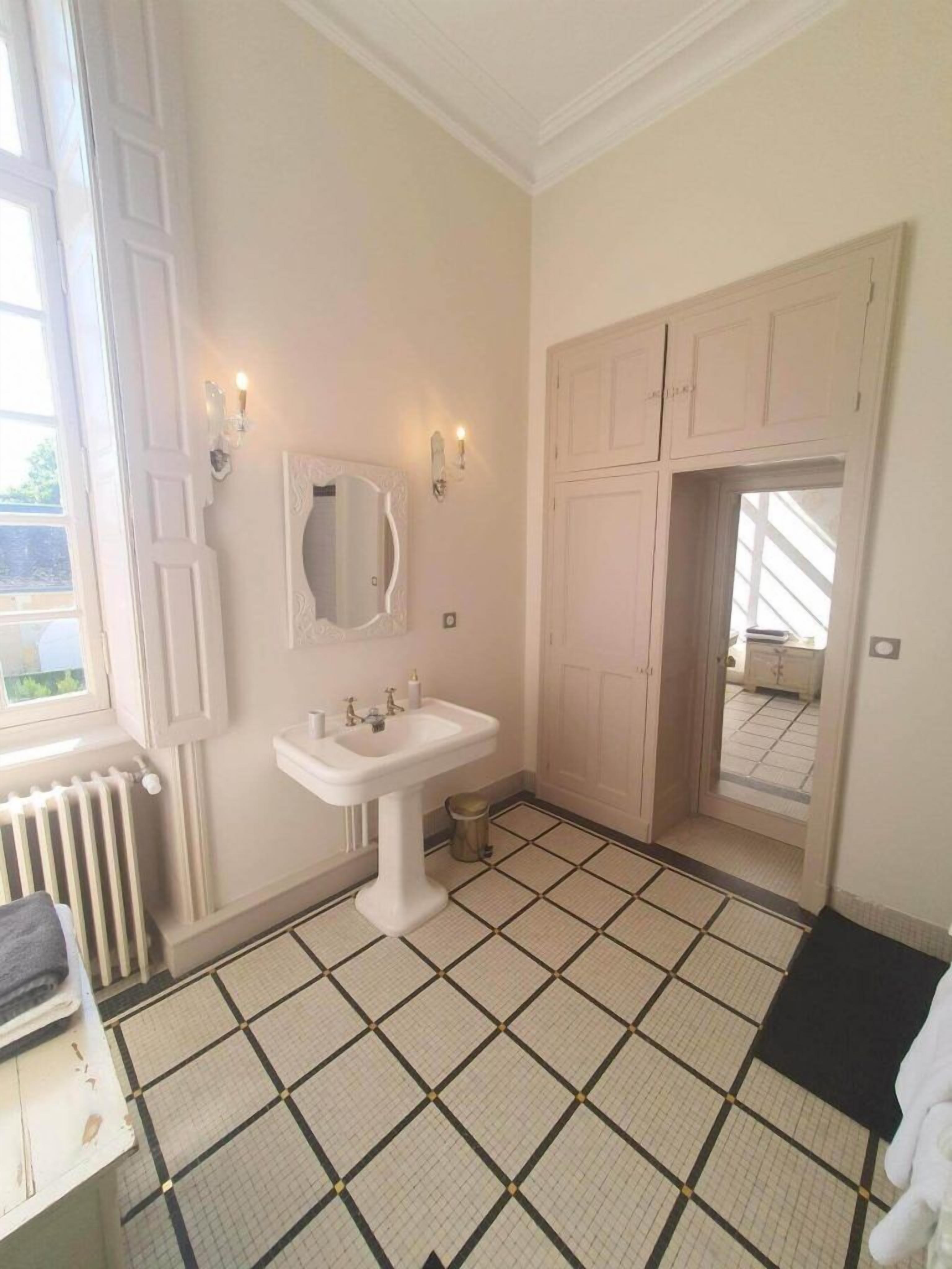 Une salle de bain avec un lavabo blanc, un miroir et deux appliques murales. On y trouve également un radiateur et une fenêtre donnant sur l’extérieur.