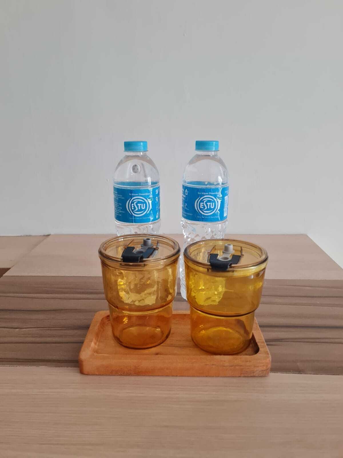 Two clear glass bottles with blue caps and labels, and two amber glass cups with black lids on a wooden tray.