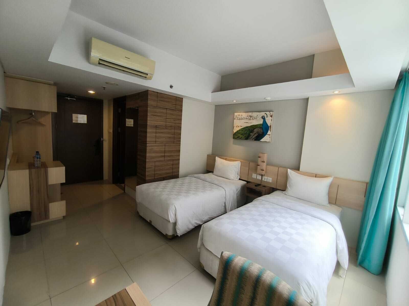 A hotel room with two beds, a wooden wardrobe, a desk, and a peacock painting on the wall.