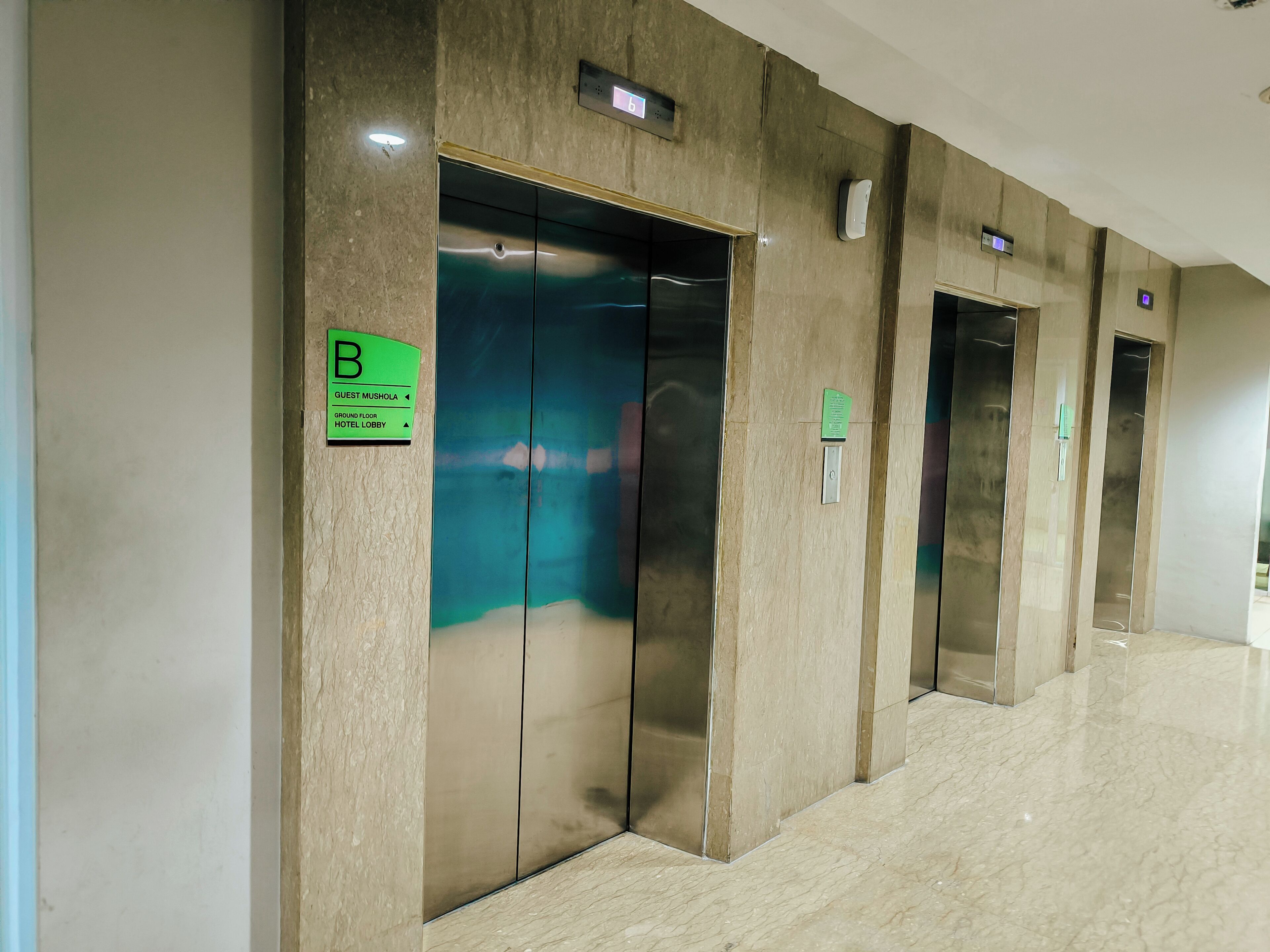 Un vestíbulo de hotel con tres ascensores, cada uno indicado con un letrero verde que muestra indicaciones hacia la sala de oración para huéspedes, la planta baja y el vestíbulo del hotel.