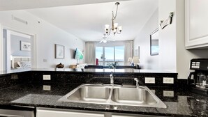 A modern kitchen with a black granite countertop, stainless steel sink, and white cabinetry. There is a ceiling fan, a chandelier, and a view of the balcony through the window.