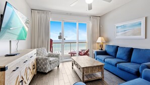 A modern living room with a large TV, a blue sofa, a wooden coffee table, and a view of the sea through the sliding glass doors.