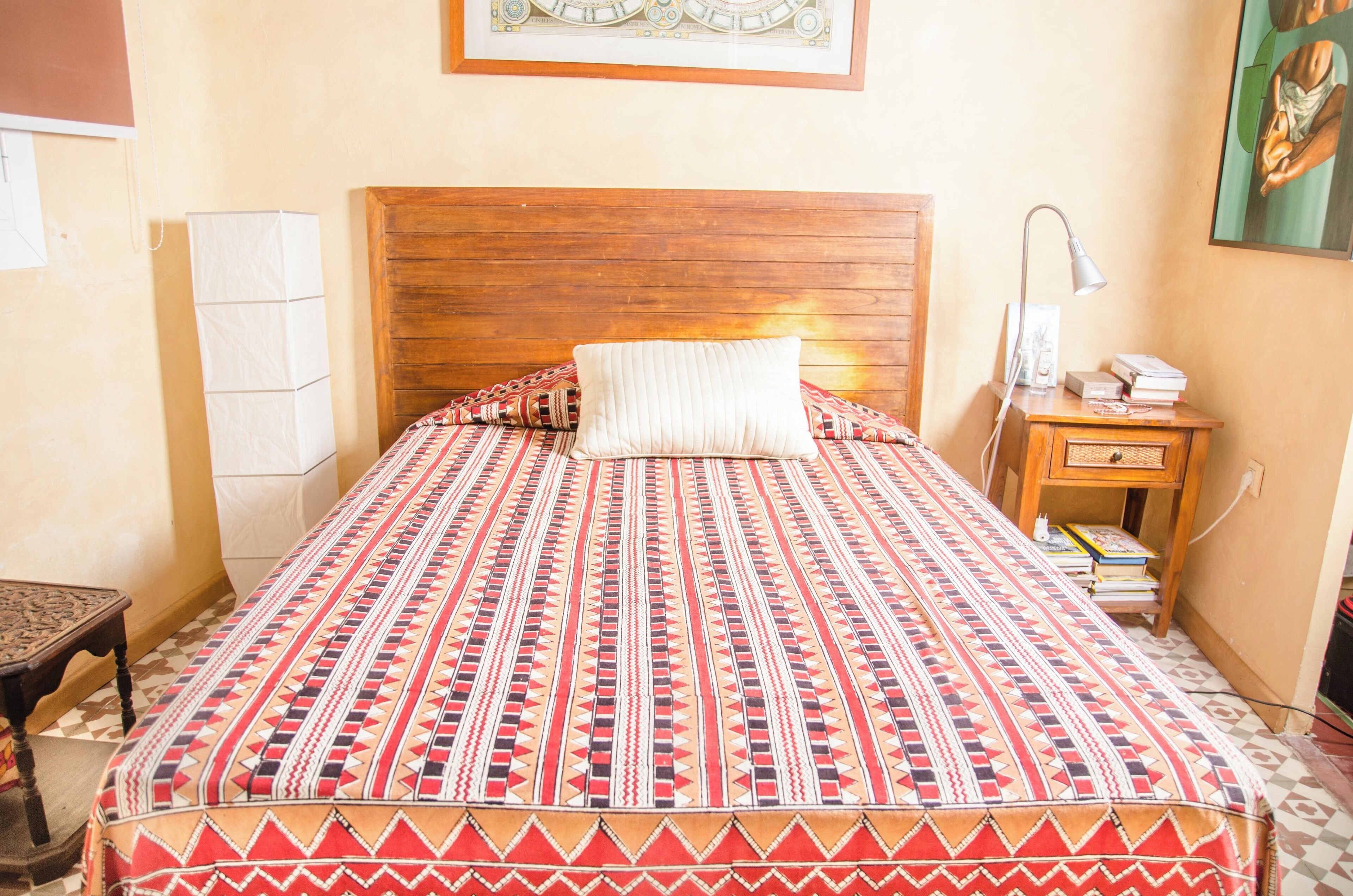 A neatly made bed with a patterned bedspread, a wooden headboard, a bedside lamp, and a nightstand with books and a phone.