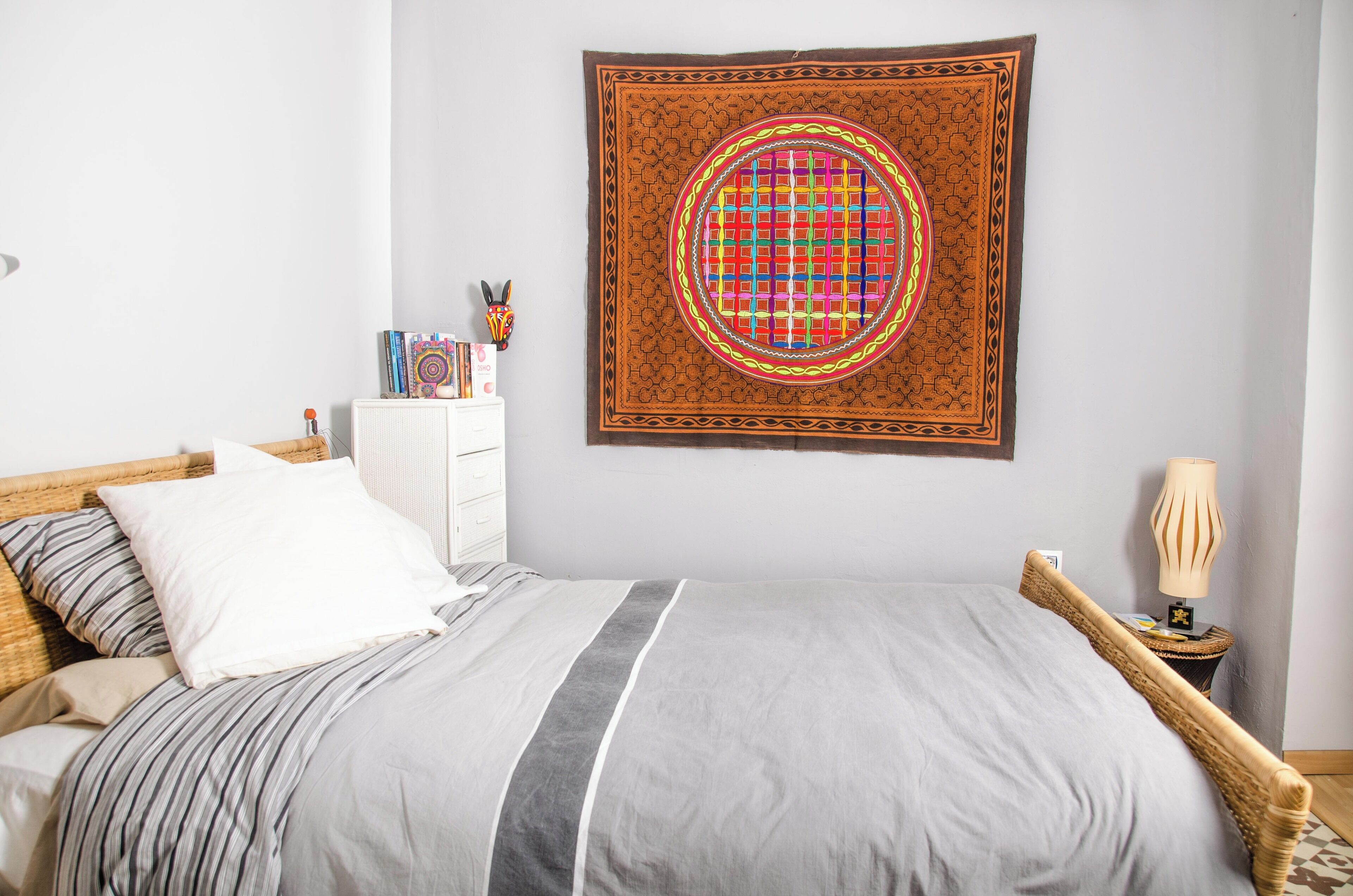 A neatly arranged bedroom with a bed, a bedside table with books, a lamp, and a tapestry on the wall.