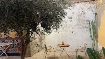 An outdoor seating area with two wicker chairs and a small table, surrounded by a tree and a cactus.