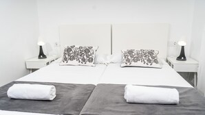 Una cama doble con colcha blanca y gris, dos almohadas con estampado floral y dos lámparas de noche.