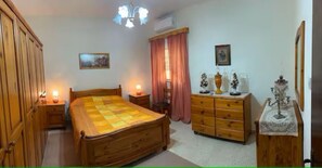 A bedroom with a wooden bed, a dresser, a nightstand, a lamp, and a window with curtains. - Lourdes House (San Gwann)
