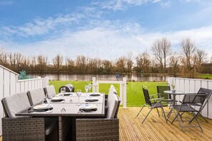 An outdoor dining area with a table set for four, surrounded by wicker chairs, overlooking a lake.
