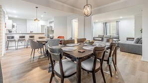 Un'ampia zona pranzo con un tavolo e sedie in legno, una cucina sullo sfondo e un salotto con divano e tende.