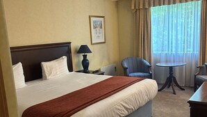 Habitación de hotel con una cama grande, una mesita de noche con lámpara, una silla, una pequeña mesa redonda y una ventana con cortinas.