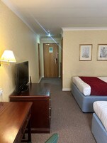 Habitación de hotel con una televisión sobre una mesa de madera, una cama con un edredón rojo y dos láminas botánicas enmarcadas en la pared.