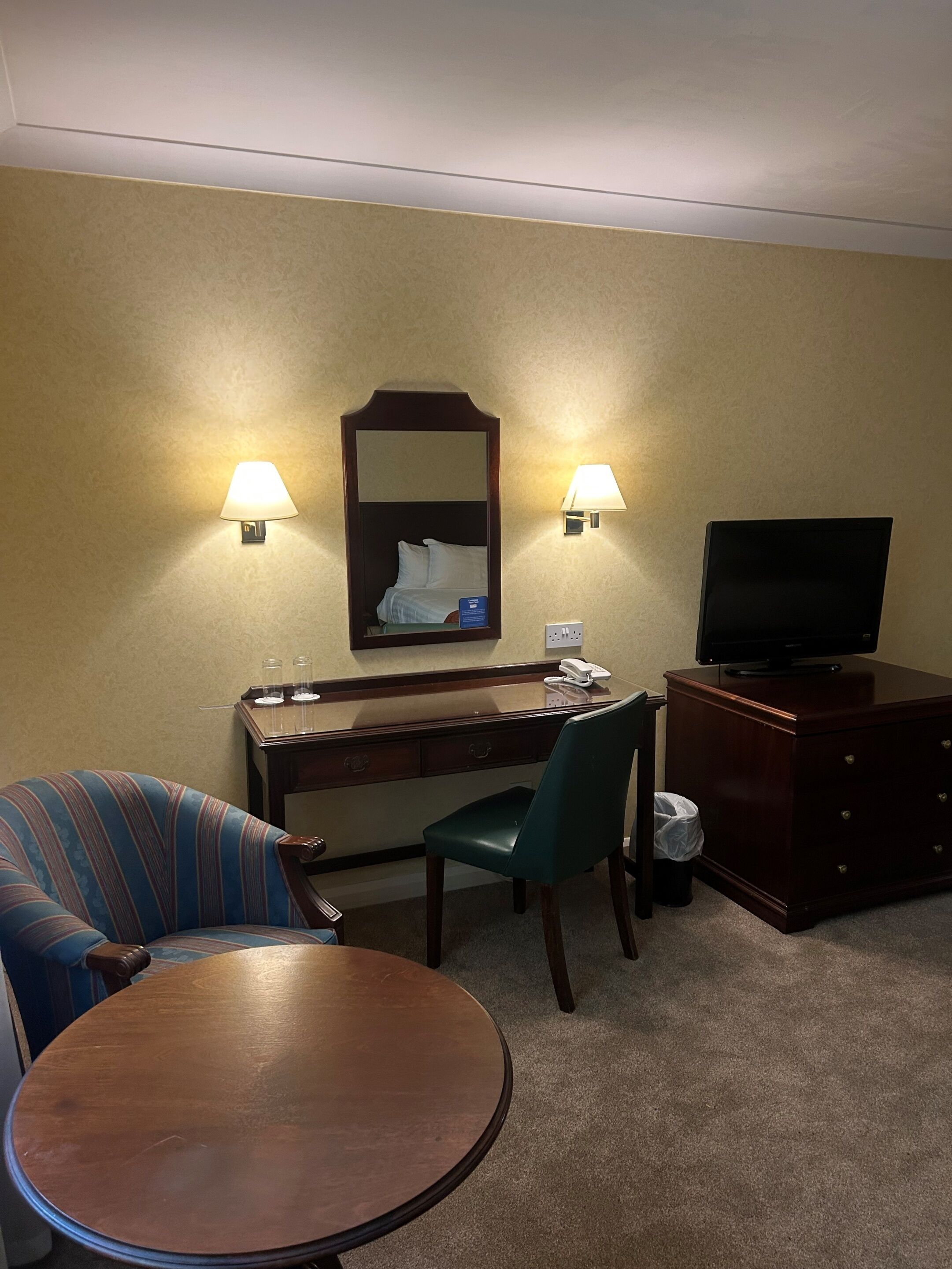 A hotel room with a bed, a desk with a mirror, a television, a chair, and a small table.