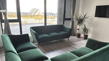 A modern living room with a large green sectional sofa, a flat-screen TV mounted on the wall, and a view of a water slide in the background.