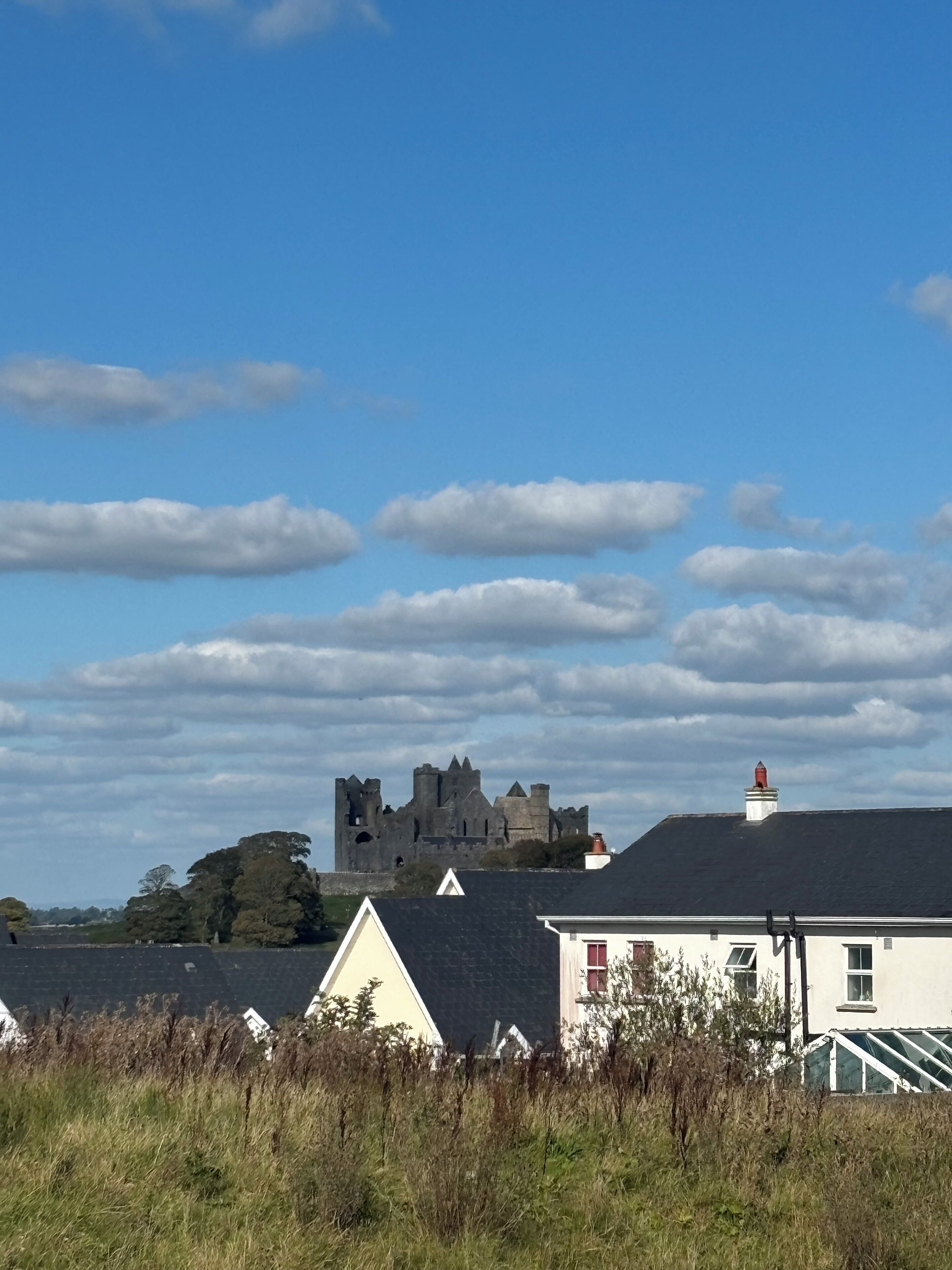 Um castelo ao fundo, com céu claro e nuvens. Em primeiro plano, casas com telhados inclinados e uma área gramada.