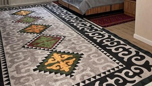 A bedroom with a bed, a nightstand, a wall-mounted artwork, a window with curtains, and a patterned rug.