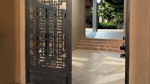 Un pasillo con una puerta decorativa, suelo de baldosas y una escalera que conduce a un espacio exterior con árboles y un banco.
