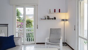 A bright living room with a white sofa, a grey armchair, a white coffee table, a bookshelf with books and decorative items, a floor lamp, and a sliding glass door leading to a balcony with plants. - Casa Bianca a Bogliasco by Wonderful Italy (Bogliasco)