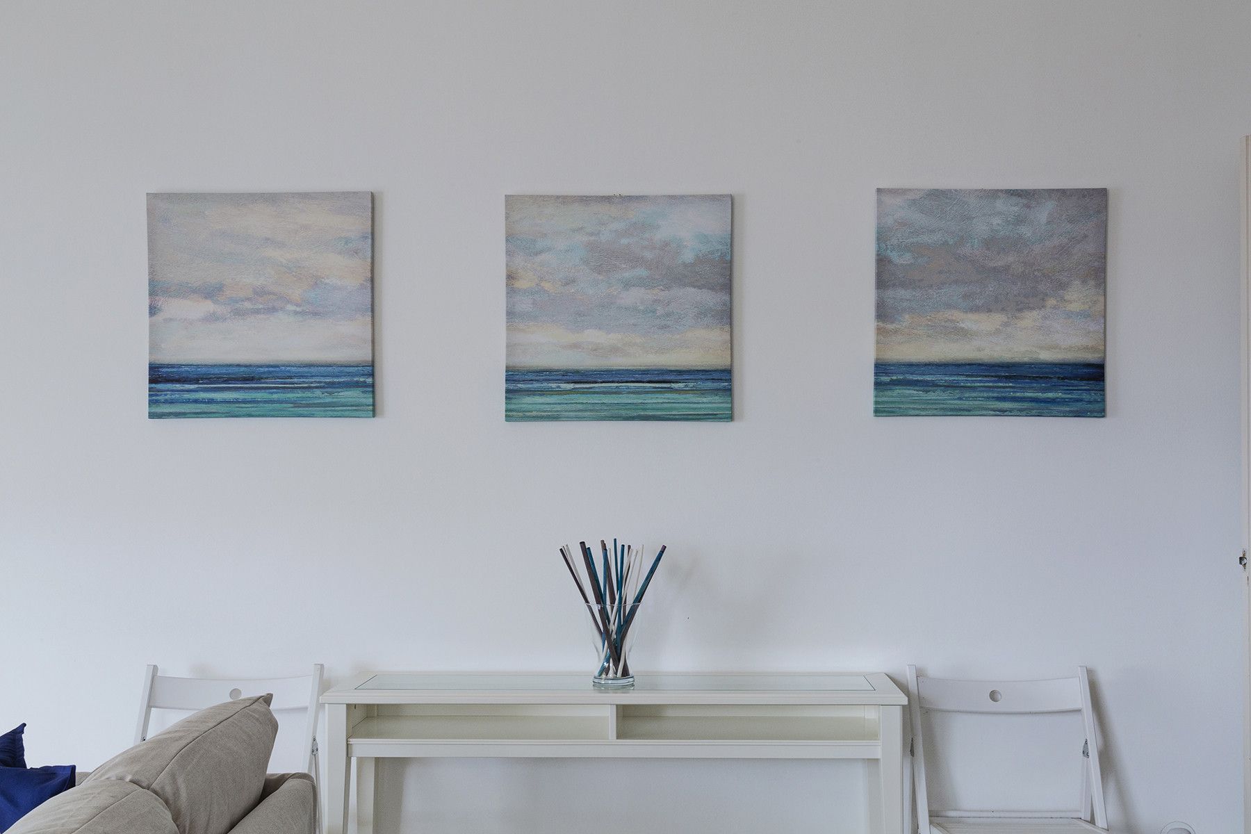 Ein Zimmer mit einer weißen Kommode, einem weißen Stuhl und drei Bildern eines bewölften Himmels über dem Meer.