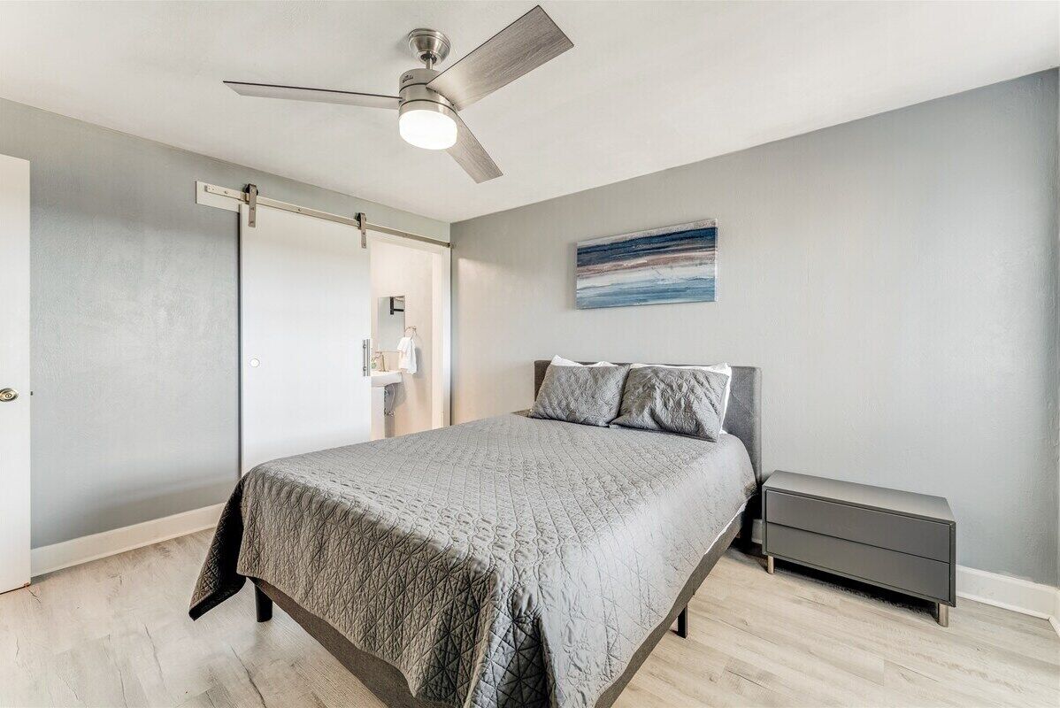 A modern bedroom with a bed, a nightstand, a ceiling fan, and a painting on the wall.