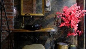 Une salle de bain avec un mur en marbre, un lavabo avec un robinet doré, un tabouret, un miroir et un vase rempli de fleurs roses.