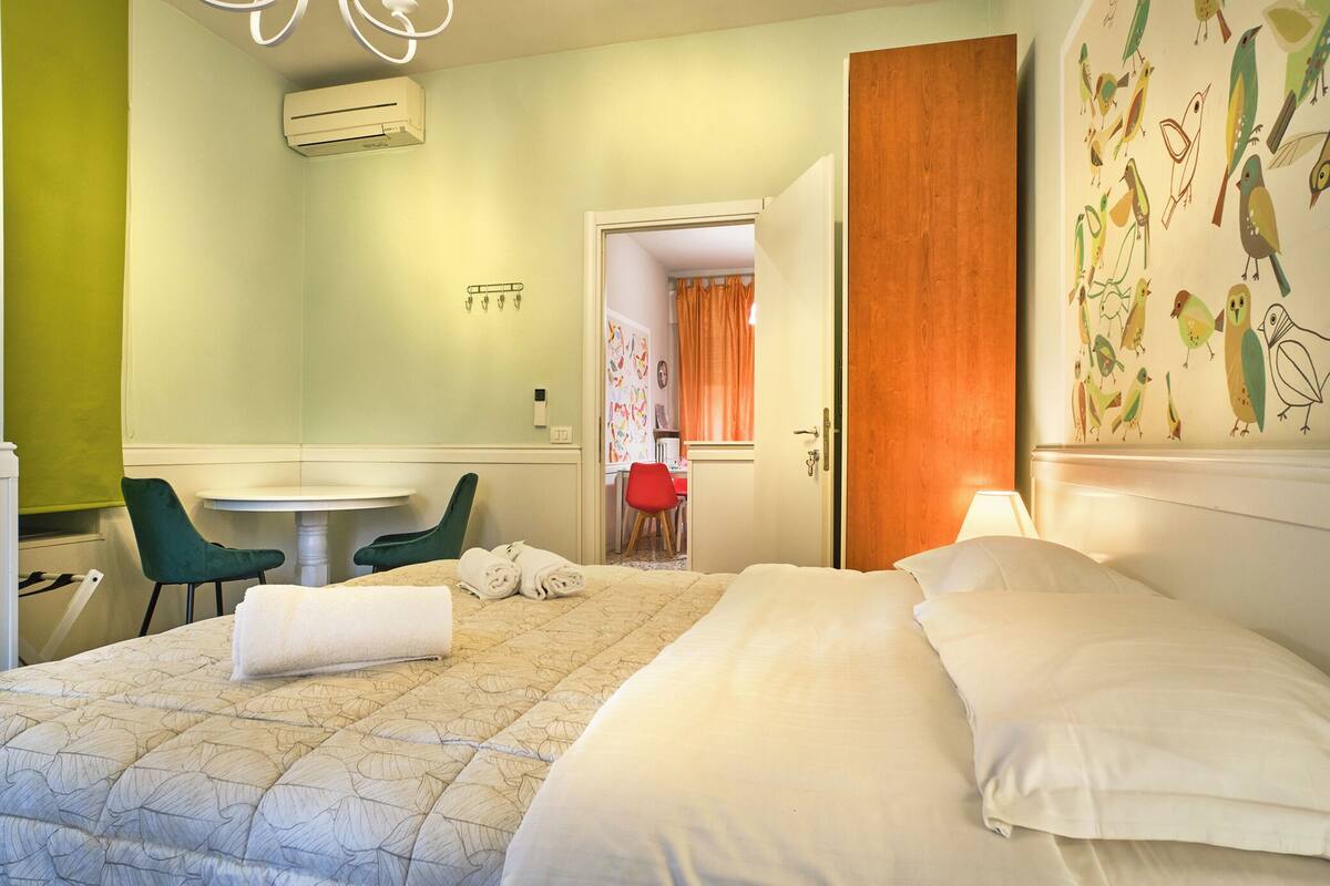 A hotel room with a bed, a round table, two chairs, a red chair, and a door leading to another room.
