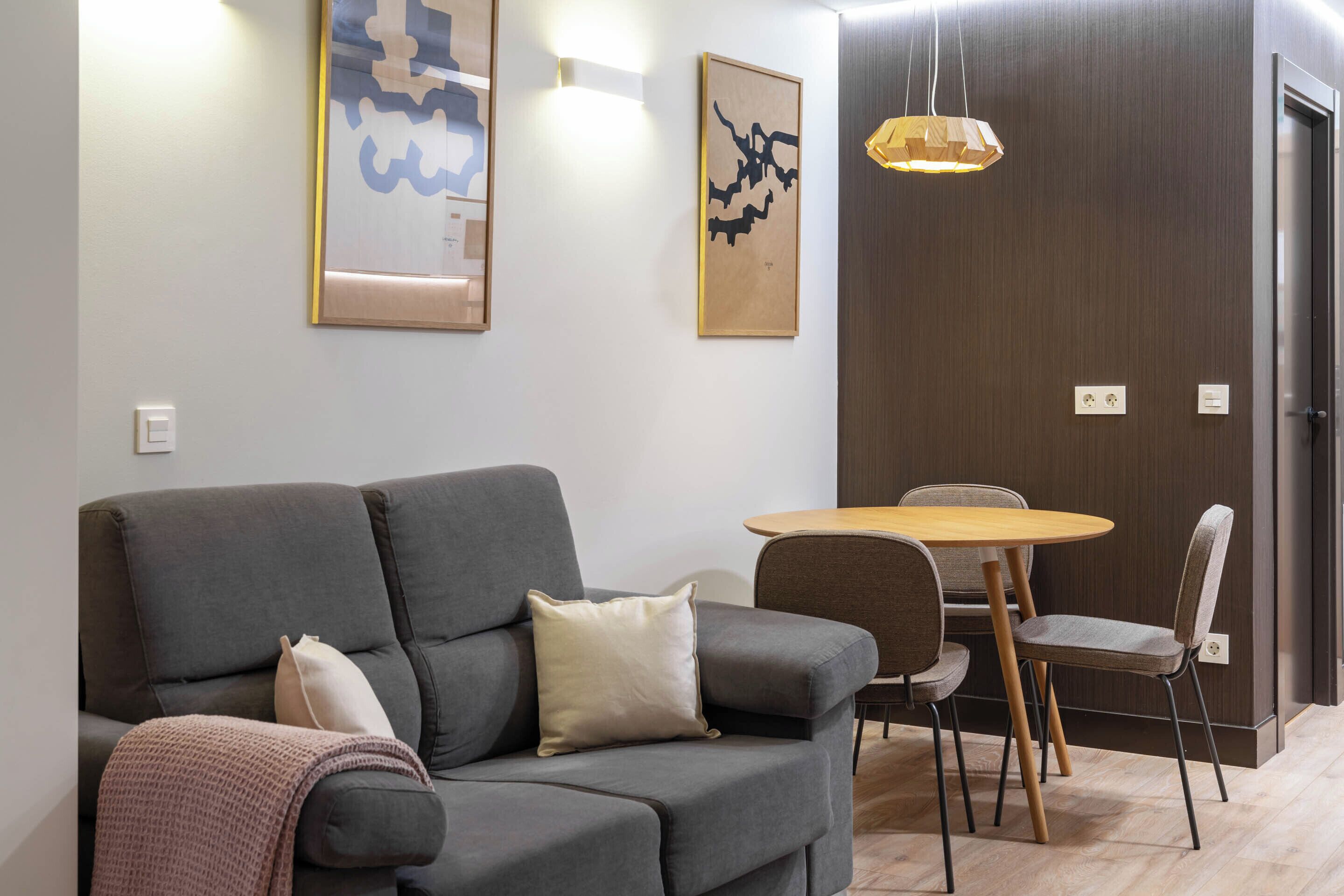 A modern living room with a grey sofa, a round wooden table, and two chairs. There are abstract paintings on the wall and a pendant light above the table.