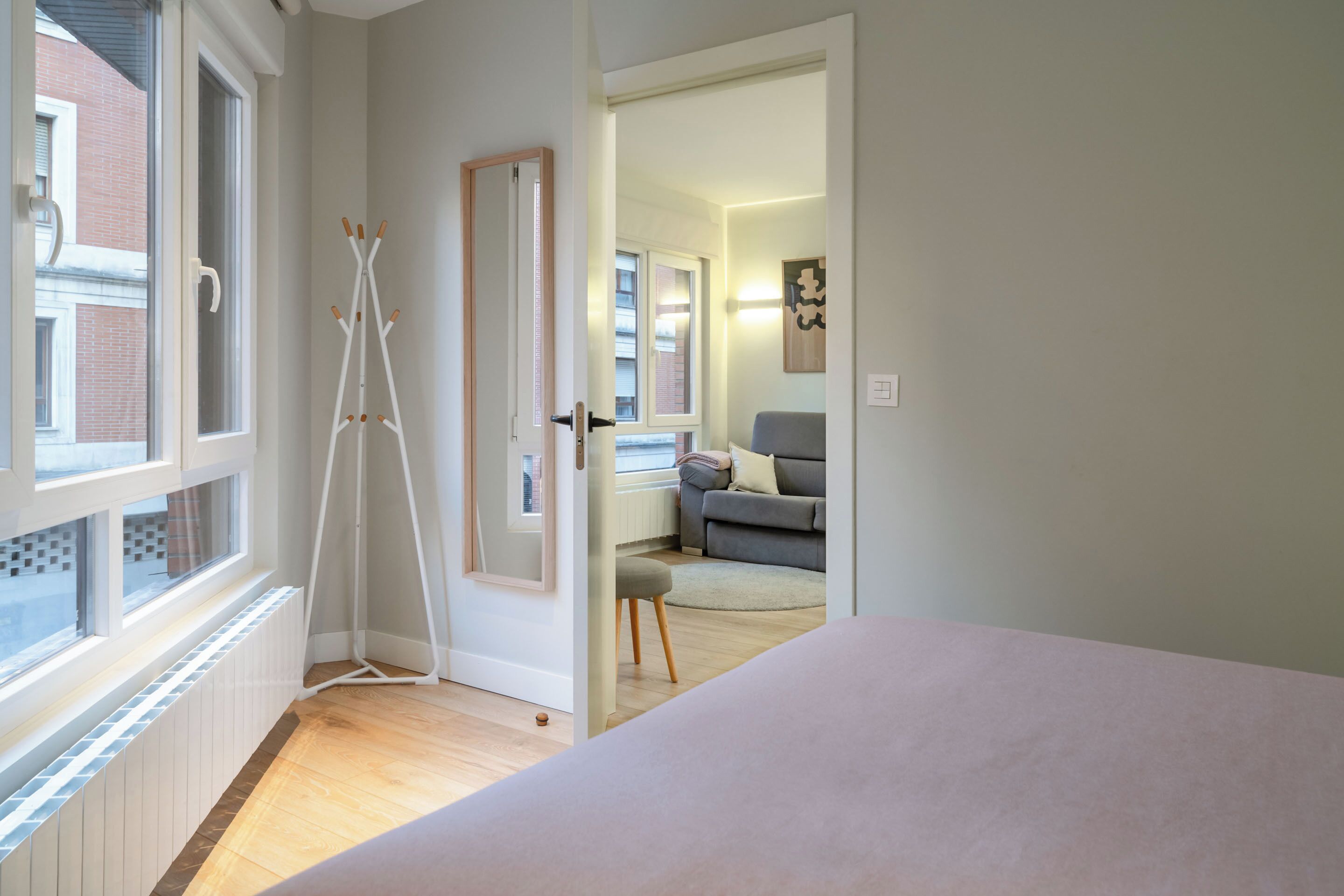 A room with a bed, a radiator, a door leading to another room, and a window with a view of a building outside.