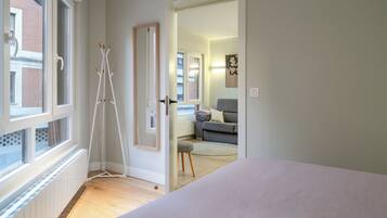 A room with a bed, a radiator, a door leading to another room, and a window with a view of a building outside.