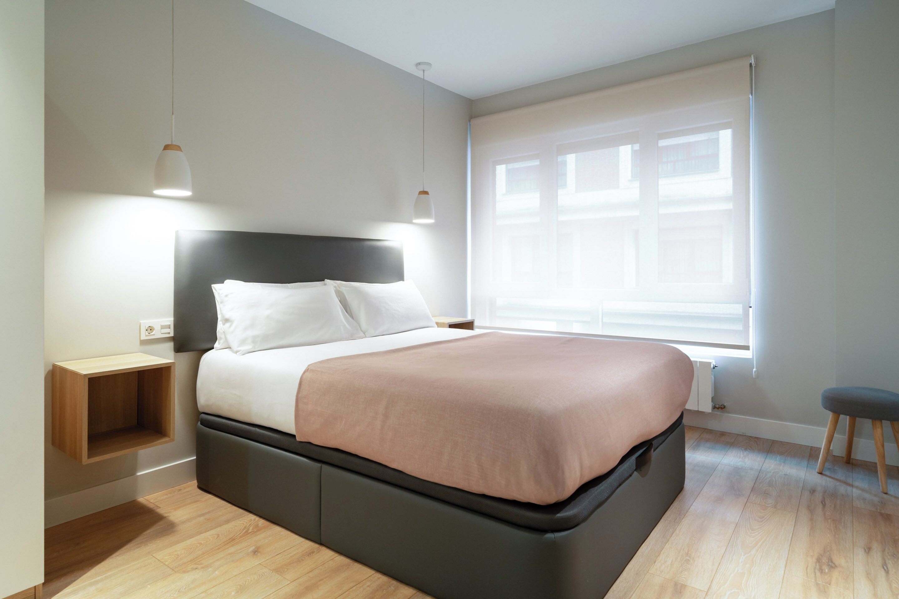 Un dormitorio moderno con cama, mesitas de noche, una ventana con persianas y un taburete.