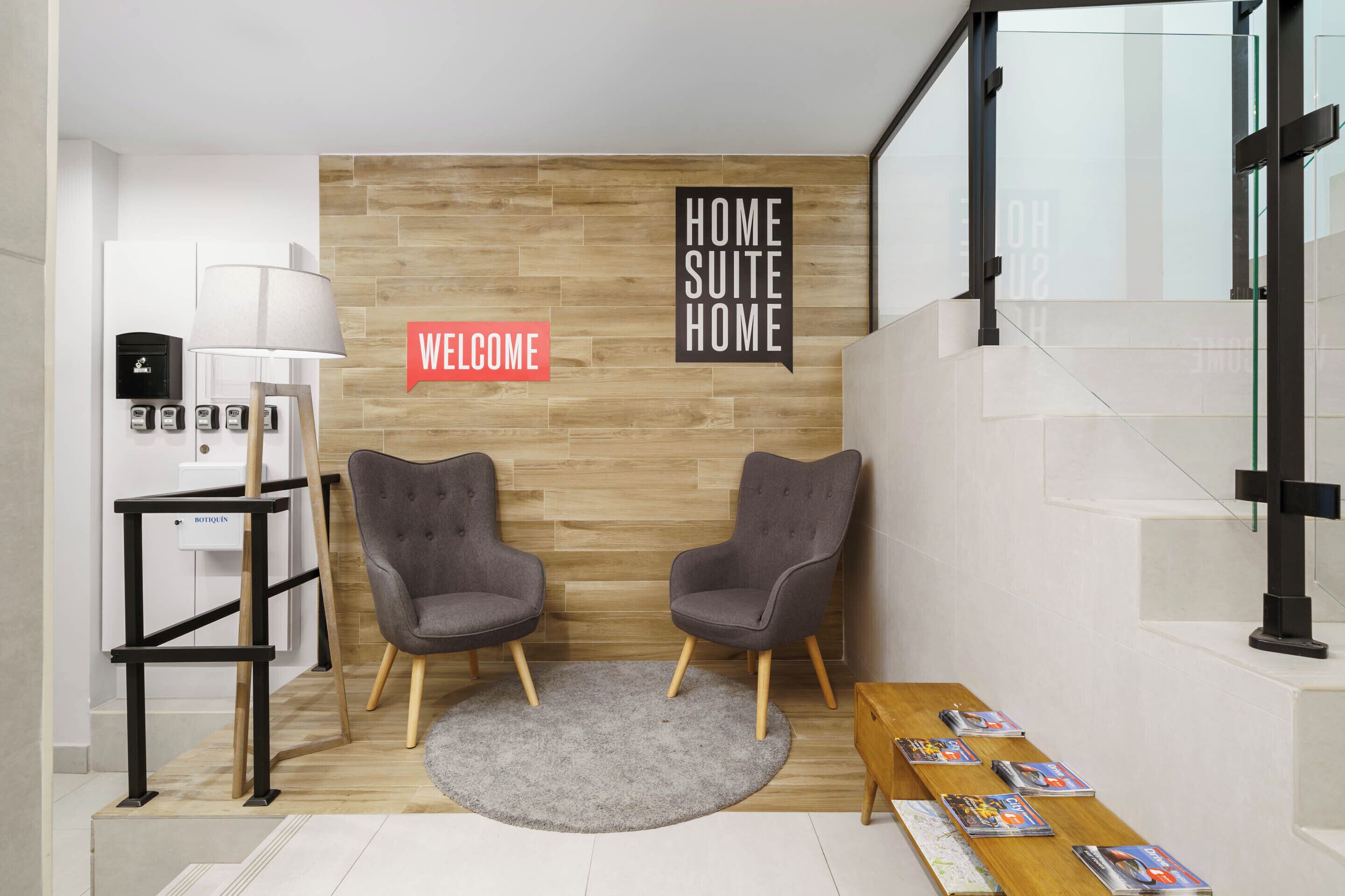 Una habitación moderna con una pared de madera, dos sillones grises, una alfombra redonda pequeña, una mesa de centro de madera con revistas, un soporte de metal negro y un cartel que dice 'BIENVENUE'.