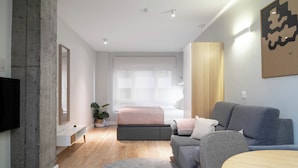A modern living room with a grey sofa, a round wooden table, a grey rug, a large window with sheer curtains, and a flat-screen TV mounted on the wall. - Limehome Bilbao Tristan de Leguizamon (Bilbao)